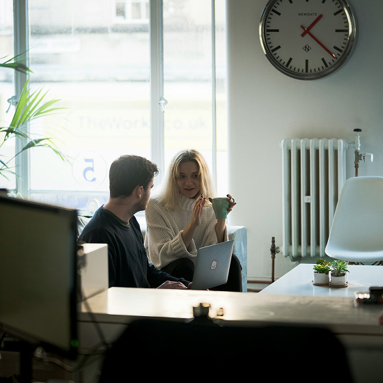 Dos personas conversando en una oficina moderna con luz natural, computadora portátil y elementos decorativos, representando colaboración y trabajo en equipo.