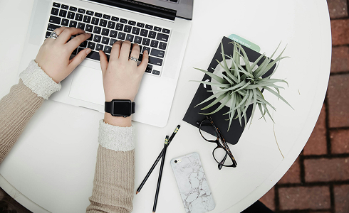 Persona trabajando en una laptop sobre una mesa blanca, con objetos de oficina como una planta, cuaderno, gafas, lápiz y teléfono móvil.